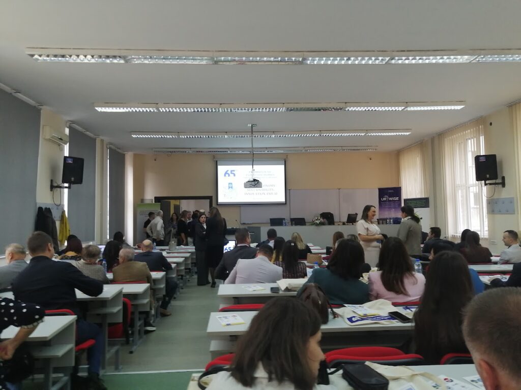 Picture of the conference room. Slide with the conference logo is displayed on the screen. Parcipants are sitting behind tables.
